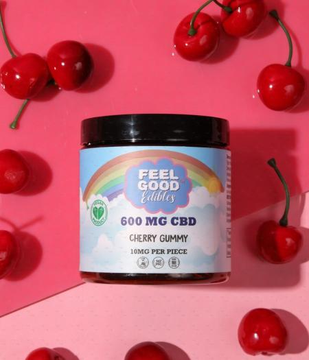 Cherry gummy container from Feel Good Edibles, featuring a colorful design and surrounded by fresh cherries on a pink background.