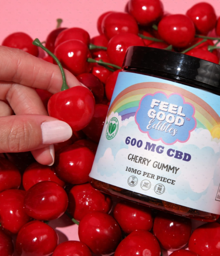 Hand holding a cherry gummy bottle surrounded by fresh cherries, showcasing the 600 mg CBD edible product.