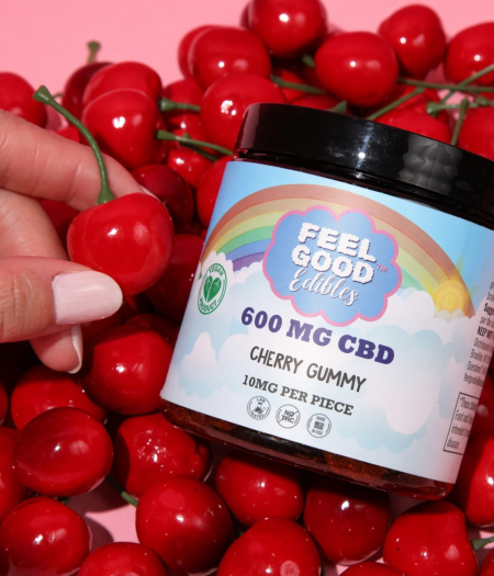 Hand holding a cherry gummy next to a jar of 600 mg CBD edibles surrounded by fresh cherries on a pink background.