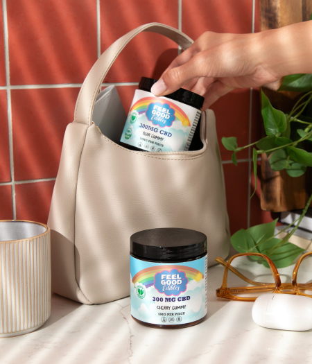 A hand retrieves a container of 300mg CBD gummies from an open beige purse on a counter with glasses and a plant.