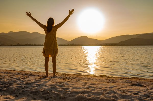 A person in a yellow dress stands by a lake at sunset, arms raised in a gesture of joy or celebration.