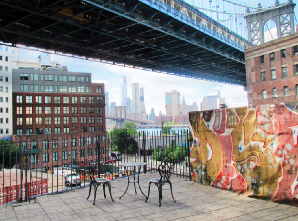 View of a Brooklyn outdoor area with a table and chairs, featuring graffiti and a skyline backdrop including the Manhattan Bridge.