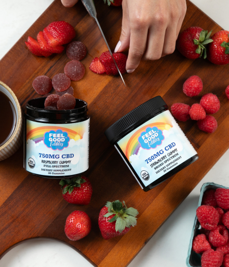 A hand slices strawberries on a wooden chopping board with jars of CBD gummies and coffee nearby.