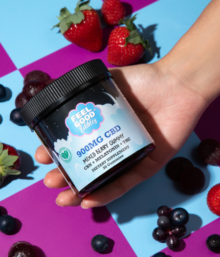 Hand holding a jar of 900mg mixed berry CBD gummies among fresh strawberries and blueberries on a colorful background.