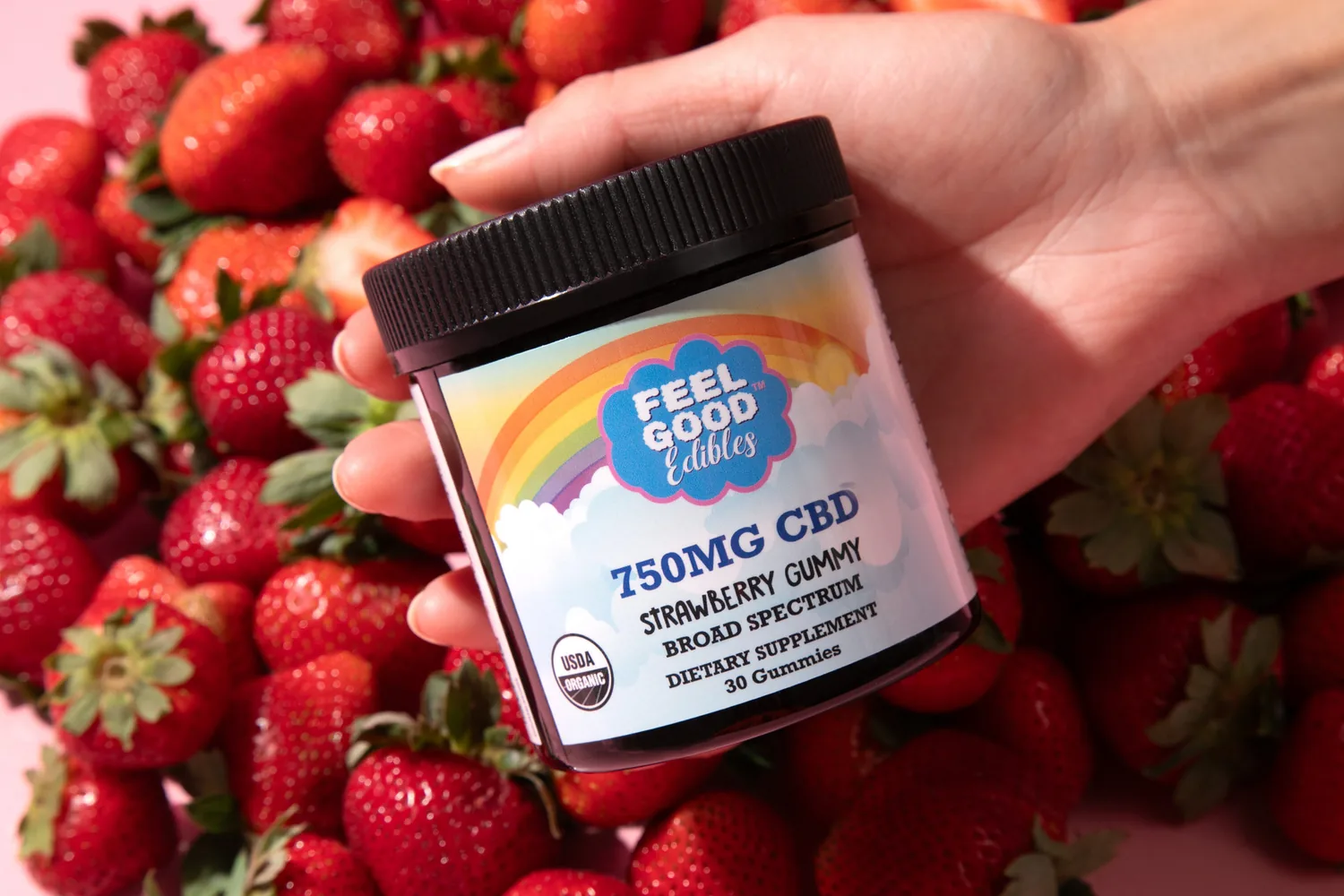 Hand holding a jar of Feel Good Edibles 750mg CBD Strawberry Gummies over fresh strawberries.